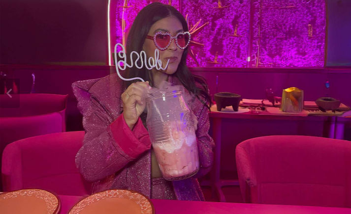 Person in a sparkly pink blazer and heart-shaped sunglasses sipping a pink milkshake from a mason jar with a curly straw in a neon-pink lounge.