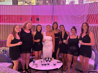 Bachelorette party group photo: eight women in cocktail dresses, bride-to-be wearing a white sash, holding cocktails in a modern indoor lounge with a pink LED crystal-bead curtain backdrop.