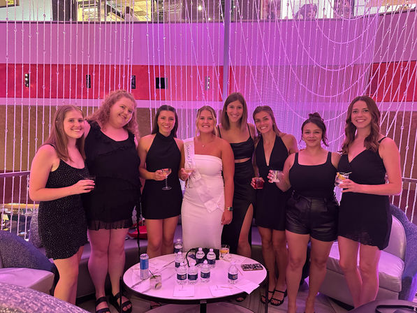 Bachelorette party group photo: eight women in cocktail dresses, bride-to-be wearing a white sash, holding cocktails in a modern indoor lounge with a pink LED crystal-bead curtain backdrop.
