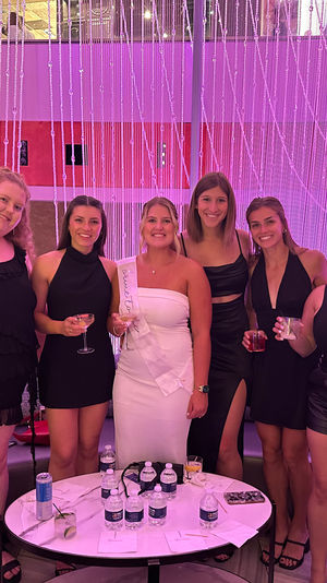 Bachelorette party group photo: eight women in cocktail dresses, bride-to-be wearing a white sash, holding cocktails in a modern indoor lounge with a pink LED crystal-bead curtain backdrop.