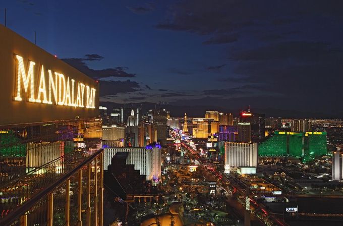 Night view of the Las Vegas Strip from a high-rise, gold hotel facade with a large illuminated sign overlooking neon-lit casinos, busy boulevard and distant city skyline at dusk.