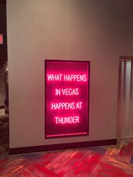 Neon pink wall sign in a Las Vegas hotel corridor reading 'What happens in Vegas...' with colorful patterned carpet and moody lighting.