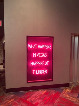 Neon pink wall sign in a Las Vegas hotel corridor reading 'What happens in Vegas...' with colorful patterned carpet and moody lighting.