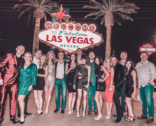 Smiling party group in festive attire posing beneath the iconic "Welcome to Fabulous Las Vegas Nevada" sign with palm trees glowing at night