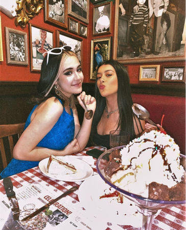 Two friends posing in a cozy retro restaurant booth with framed photos on the red wall, holding spoons over a slice of cake and a giant ice cream sundae on a red-and-white checkered tablecloth.