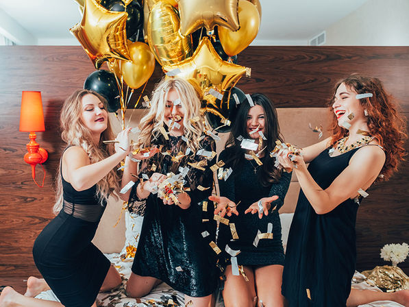 Four friends in black dresses toss gold and silver confetti on a bed under gold star balloons in a chic hotel-room setting, playful festive celebration (party, bachelorette or birthday vibe)