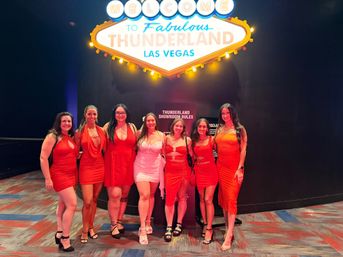 Seven women in red dresses posing beneath a bright Las Vegas marquee sign inside a casino showroom