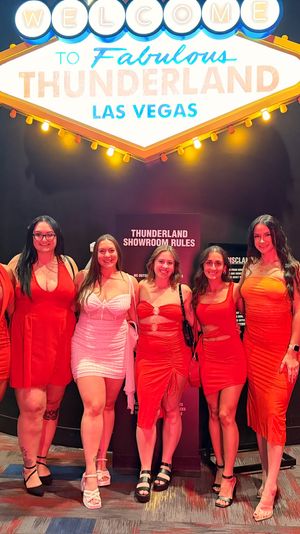 Seven women in red dresses posing beneath a bright Las Vegas marquee sign inside a casino showroom