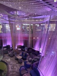 Indoor atrium lounge with purple LED lighting and glittering crystal bead curtains, curved sofas and round tables on a marbled floor seen from an upper level