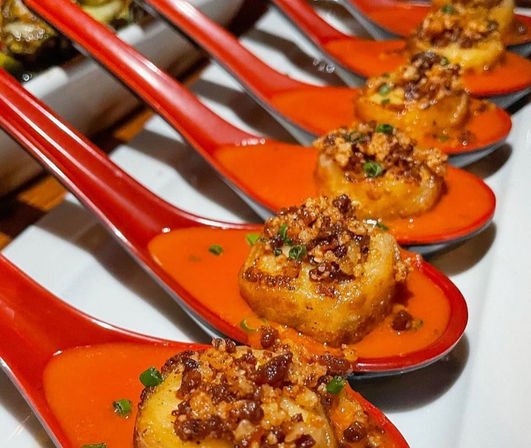 Row of restaurant-style seared scallop appetizers on red tasting spoons, each perched in a bright red-orange sauce and topped with crispy breadcrumbs and chopped chives on a white platter.
