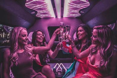 Four women toasting champagne inside a neon pink-purple stretch limousine during an urban nightlife celebration.