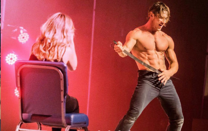 Shirtless male dancer brandishing a sword on a red-lit stage, performing energetically for a seated spectator with long blonde hair.