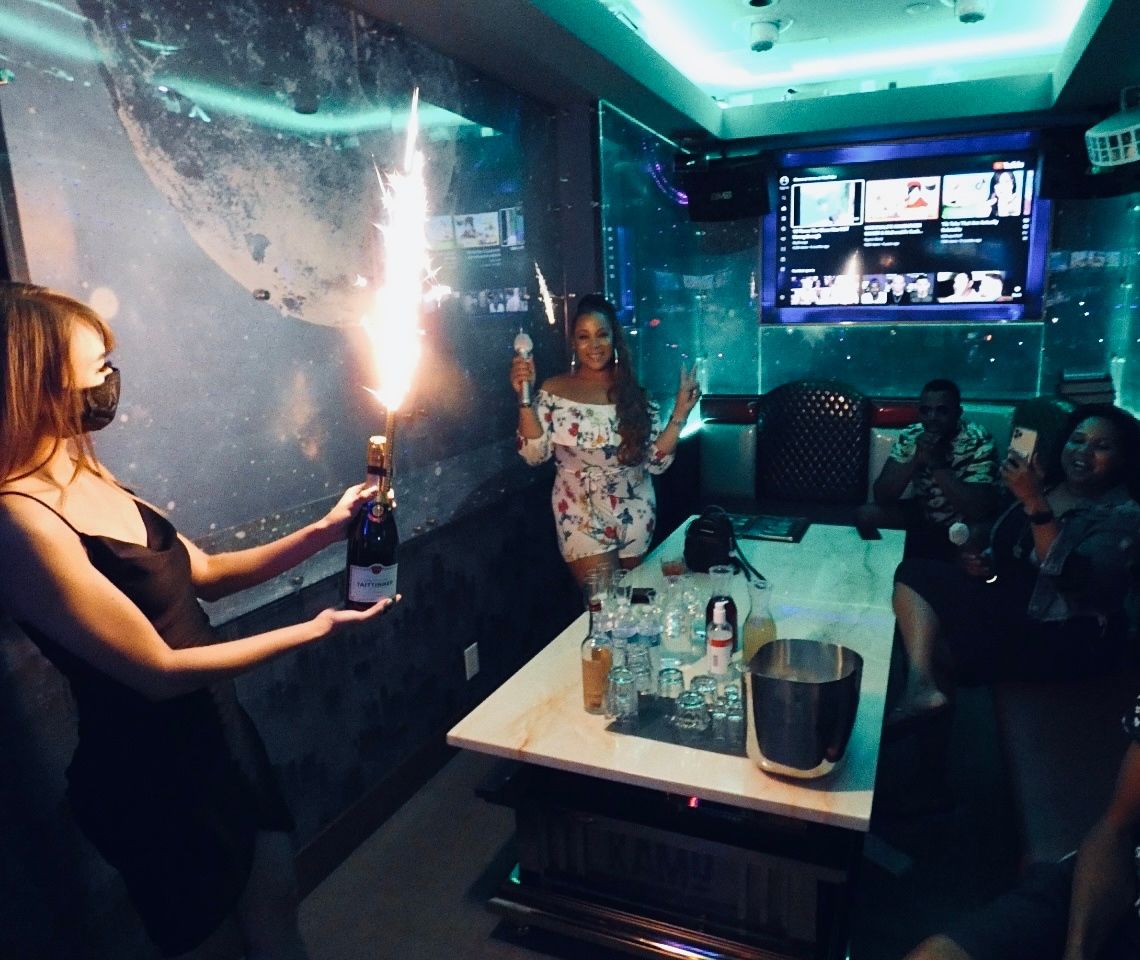 Neon-lit private karaoke room celebration with bottle service sparkler, woman posing with microphone by table of drinks as guests record on phones