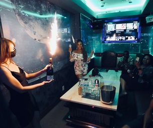 Neon-lit private karaoke room celebration with bottle service sparkler, woman posing with microphone by table of drinks as guests record on phones