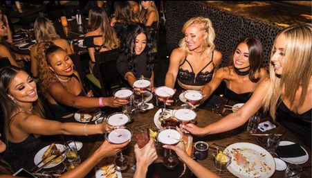 Group of friends toasting cocktails around a lively restaurant table with plates of food, capturing a girls' night out vibe.
