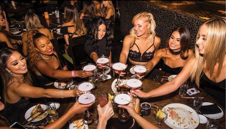Group of friends toasting cocktails around a lively restaurant table with plates of food, capturing a girls' night out vibe.