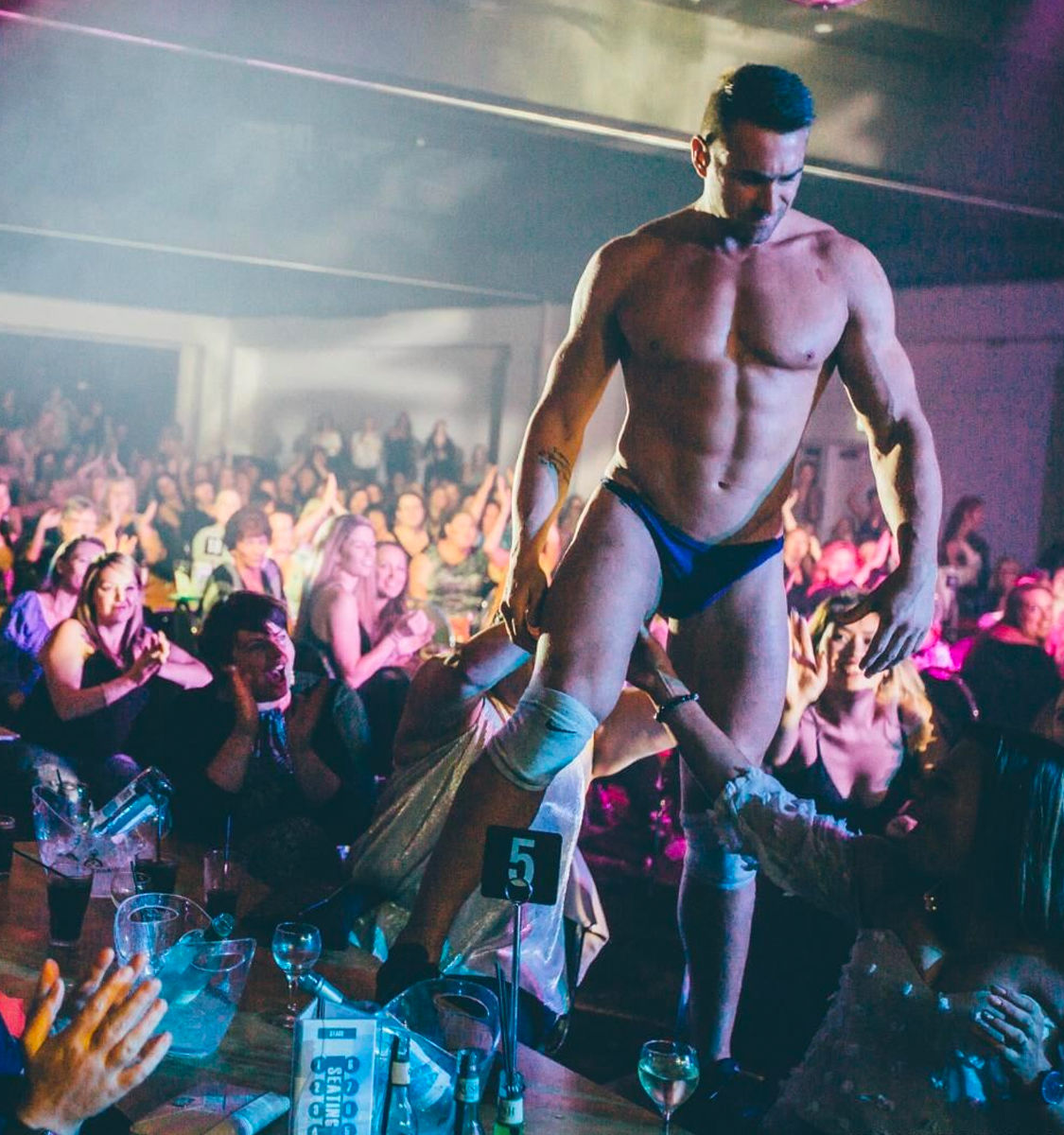 Muscular male performer in blue briefs entertains a cheering nightclub crowd, walking on tables and reaching for audience hands under colorful stage lights.