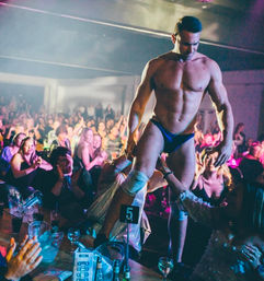 Muscular male performer in blue briefs entertains a cheering nightclub crowd, walking on tables and reaching for audience hands under colorful stage lights.