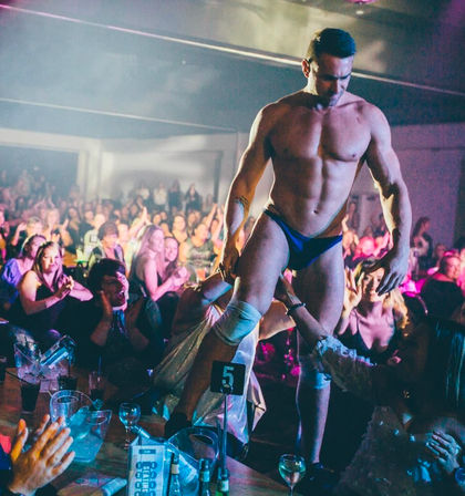 Muscular male performer in blue briefs entertains a cheering nightclub crowd, walking on tables and reaching for audience hands under colorful stage lights.