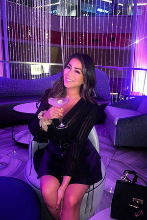 Smiling woman in a black dress holding a coupe cocktail in a purple-lit upscale lounge with shimmering beaded curtains, modern sofas and marble-pattern floor.
