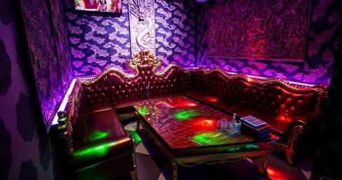 Vibrant purple-lit VIP karaoke room with tufted red leather sectional, gold dragon carvings, marble coffee table with glasses and remote, wall-mounted TV and colorful disco lights.