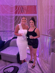 Two smiling women holding cocktails in a pink-purple lit lounge; one in a white dress with a bride-to-be sash and the other in a black top and shorts, posing in front of a beaded string curtain.