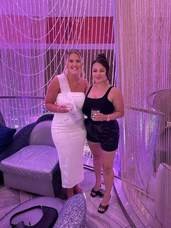 Two smiling women holding cocktails in a pink-purple lit lounge; one in a white dress with a bride-to-be sash and the other in a black top and shorts, posing in front of a beaded string curtain.