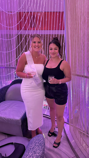 Two smiling women holding cocktails in a pink-purple lit lounge; one in a white dress with a bride-to-be sash and the other in a black top and shorts, posing in front of a beaded string curtain.