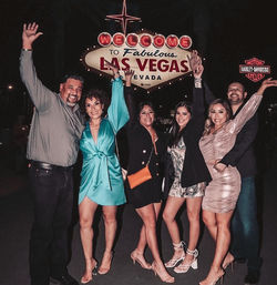 Six friends dressed up at night, smiling and raising their arms in front of the illuminated Welcome to Fabulous Las Vegas Nevada sign on the Strip.