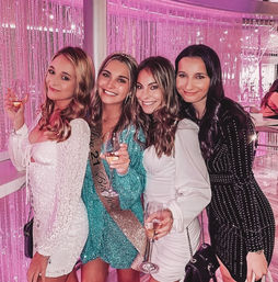 Four smiling women in sparkly cocktail dresses holding champagne glasses in a pink-lit lounge — one wearing a birthday sash and tiara for a glam night out.