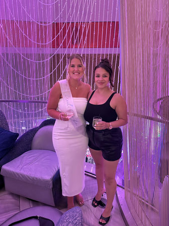 Two smiling friends at a pink-lit lounge with a beaded curtain backdrop — one in a white dress and sash holding a wine glass, the other in a black top and leather shorts holding a cocktail.