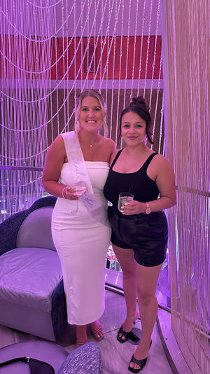 Two smiling friends at a pink-lit lounge with a beaded curtain backdrop — one in a white dress and sash holding a wine glass, the other in a black top and leather shorts holding a cocktail.
