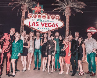 Cheerful group of well-dressed people posing at night in front of the Welcome to Fabulous Las Vegas Nevada sign, framed by palm trees and neon lights.