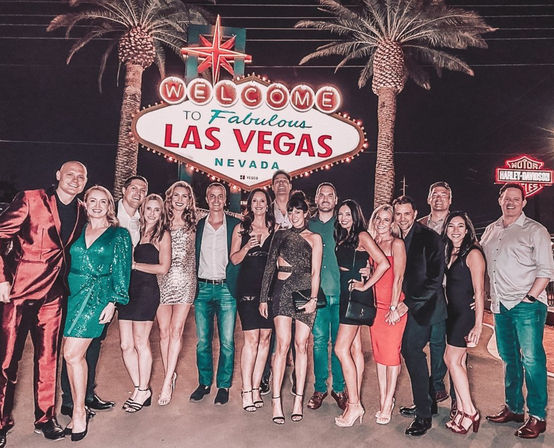 Group of friends in stylish evening outfits posing at night beneath the illuminated Welcome to Fabulous Las Vegas Nevada sign framed by palm trees