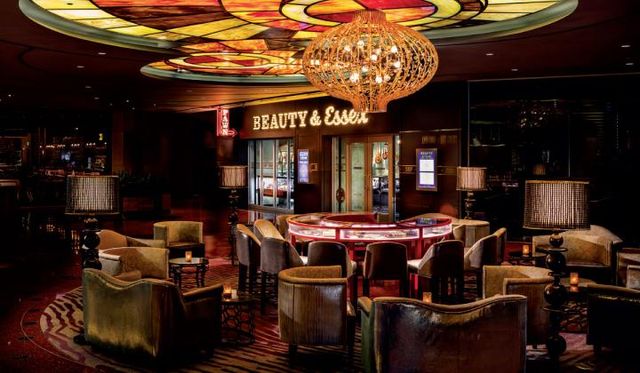 Upscale lounge and cocktail bar interior with plush velvet chairs around a circular table, stained-glass ceiling panels, ornate chandelier and warm ambient lighting.