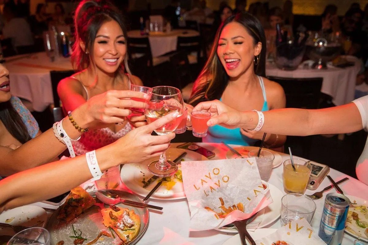 Friends clinking wine and shot glasses over shared plates at a lively restaurant dinner — girls' night out celebration.
