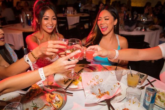 Friends clinking wine and shot glasses over shared plates at a lively restaurant dinner — girls' night out celebration.