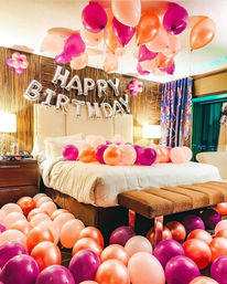 Balloon-filled birthday bedroom surprise with silver "HAPPY BIRTHDAY" foil letters and dozens of pink, peach, and magenta balloons scattered on the bed, floor, and ceiling in a cozy hotel-style room.