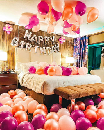 Balloon-filled birthday bedroom surprise with silver "HAPPY BIRTHDAY" foil letters and dozens of pink, peach, and magenta balloons scattered on the bed, floor, and ceiling in a cozy hotel-style room.