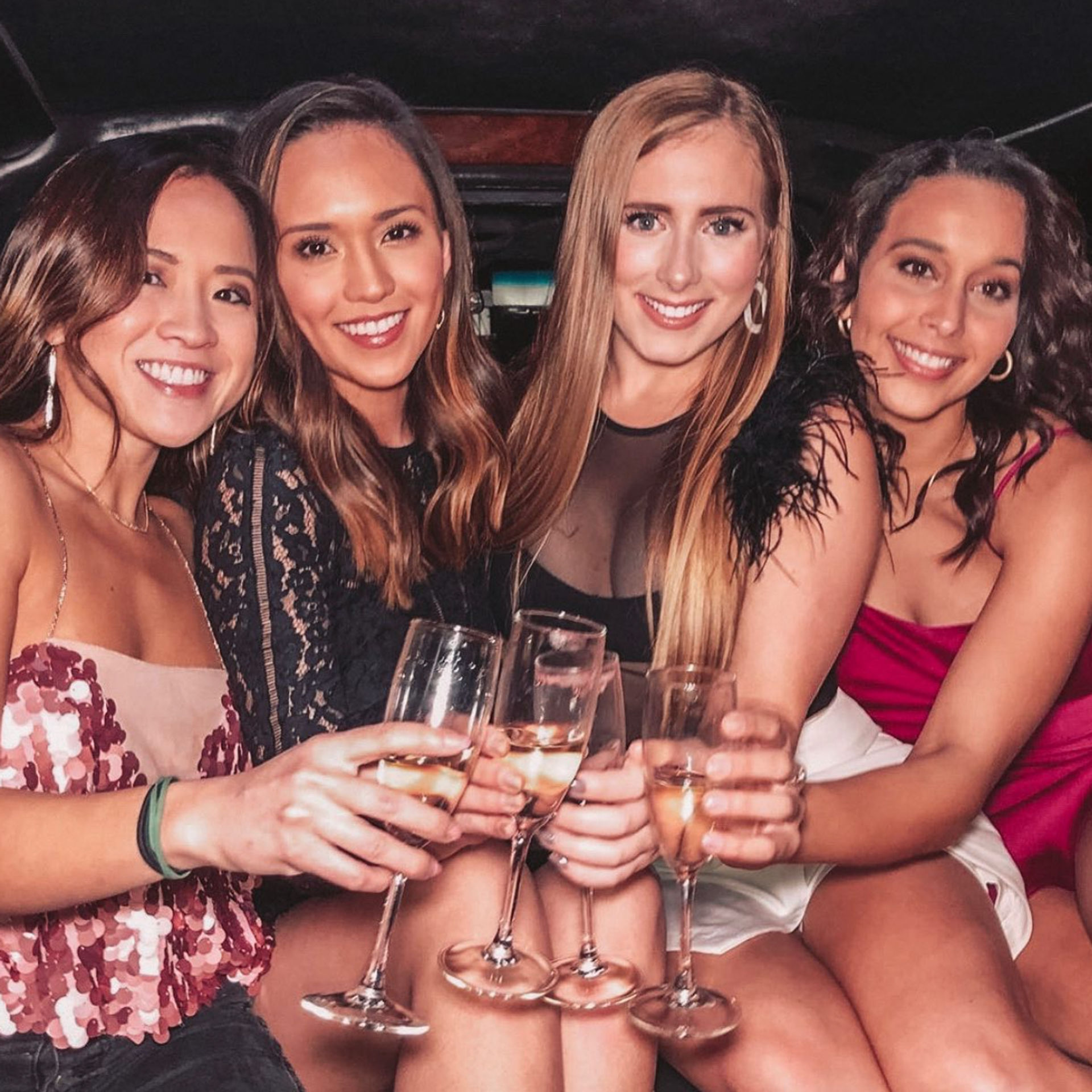 Four smiling women in cocktail dresses clinking champagne flutes inside a limo for a festive girls’ night out — city nightlife celebration.