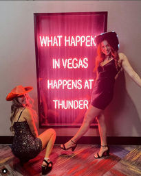 Party-ready duo in black dresses and cowgirl hats pose by a pink neon sign reading "WHAT HAPPENS IN VEGAS", capturing a playful Las Vegas nightlife vibe.