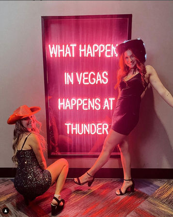 Party-ready duo in black dresses and cowgirl hats pose by a pink neon sign reading "WHAT HAPPENS IN VEGAS", capturing a playful Las Vegas nightlife vibe.
