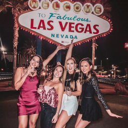 Four women in party dresses holding champagne, posing at night in front of the iconic Welcome to Fabulous Las Vegas Nevada sign with palm trees and city lights