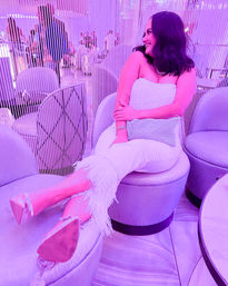 Smiling woman in a white strapless outfit with feather-trimmed trousers reclining on a round ottoman in a pink-purple neon-lit lounge with vertical beaded curtain and modern seating