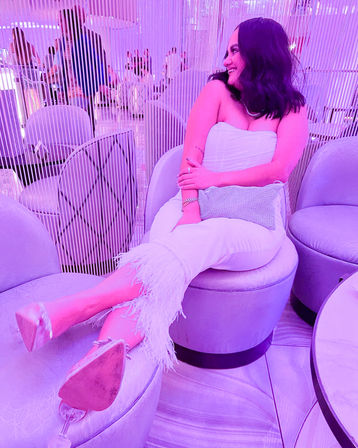 Smiling woman in a white strapless outfit with feather-trimmed trousers reclining on a round ottoman in a pink-purple neon-lit lounge with vertical beaded curtain and modern seating