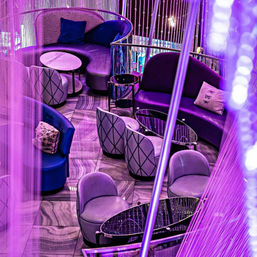 Purple-lit modern lounge with curved velvet sofas, patterned club chairs, marble-top tables and beaded curtain partitions — chic city nightlife seating.