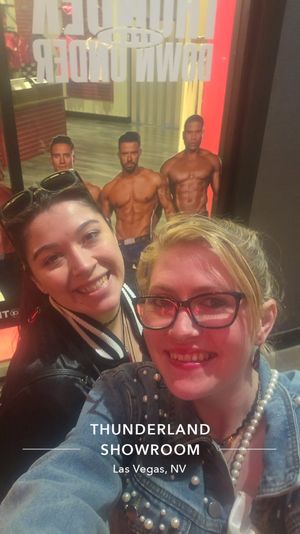Two smiling women take a selfie in a Las Vegas showroom window with cardboard cutouts of shirtless muscular men and red neon lighting