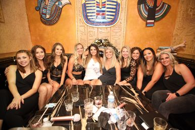 Smiling group of 11 women in party dresses posing in an Egyptian‑themed lounge booth around a marble table with drinks, microphones and a bride‑to‑be wearing a white sash.