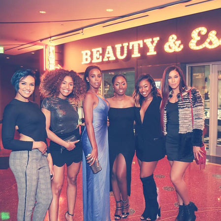 Six stylish women in eveningwear posing inside a lit venue under a glowing marquee sign, smiling for a night-out group photo