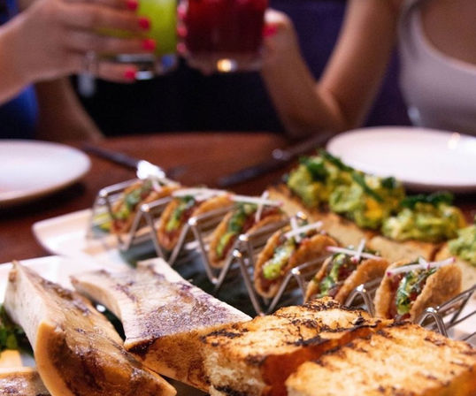 Restaurant appetizer spread: mini tacos in a metal rack, roasted bone marrow with grilled crostini and avocado toast, diners clinking colorful cocktails.